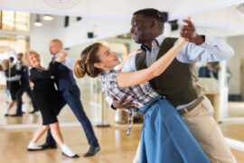 A well-dressed duo practicing ballroom dancing in a dance hall with another duo dancing behind them.