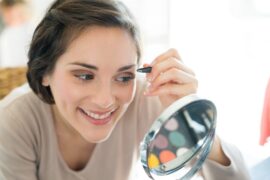 A close-up of a woman looking in a small, round, mirror stand as she holds a pair of tweezers, plucking her eyebrows.