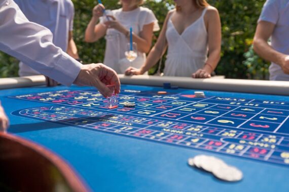 Close-up of a blue roulette table surrounded by guests at an outdoor daytime party. The dealer moves the roulette marker.