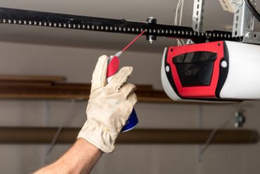 A person wearing gloves applies oil in a spray can to the chain on a garage door opener. The opener is white and red.