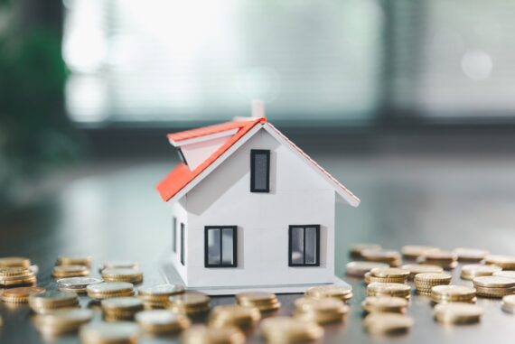 A white model house with a red roof surrounded by stacks of coins on a black surface in front of a window.