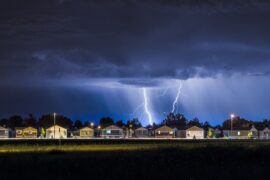 A neighborhood is full of houses in the distance, sitting underneath a thunder cloud with lightning bolts coming down.