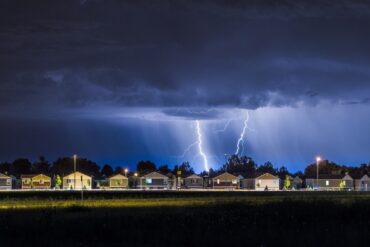 A neighborhood is full of houses in the distance, sitting underneath a thunder cloud with lightning bolts coming down.