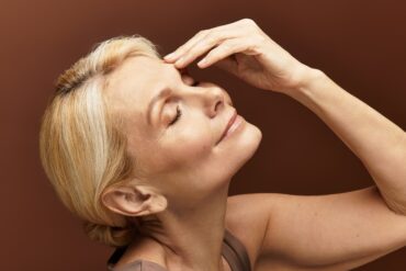 A portrait of an older woman with blonde hair and some gray streaks. She holds her hand gently to her brow and closes her eyes.