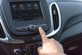 A close-up of a vehicle's stereo. There are icons on the display, and someone's finger presses a button beneath the screen.
