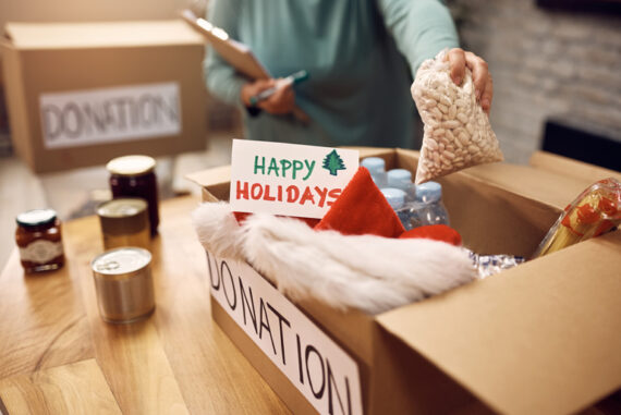 A person filling a donation box with everyday essentials and a note that reads "Happy Holidays." They are holding a clipboard.
