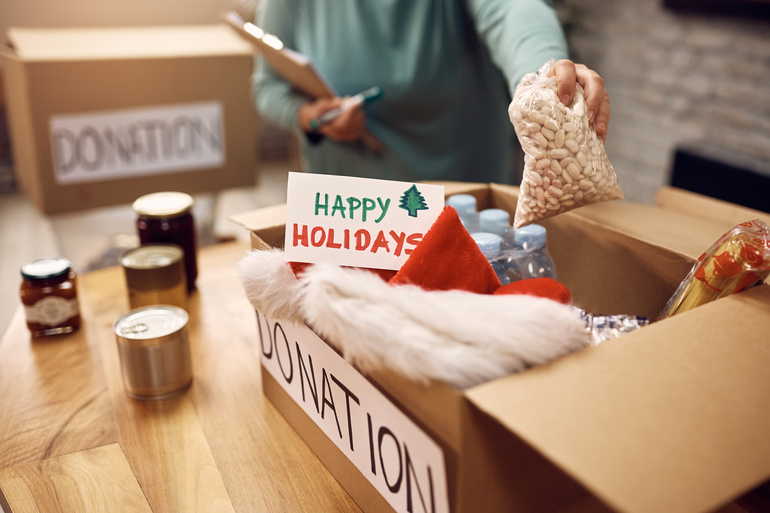 A person filling a donation box with everyday essentials and a note that reads "Happy Holidays." They are holding a clipboard.