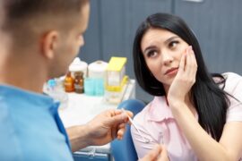 A woman in a pink shirt sits in a dental chair, looking at a dental professional holding her left cheek.