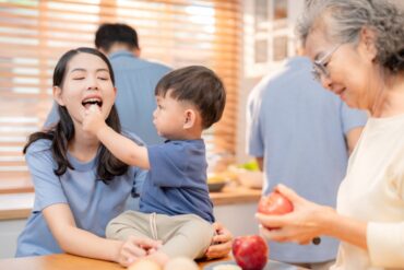 A multi-generational Asian family sits in the kitchen; the grandmother is cutting fruit, and the child feeds it to the mom.