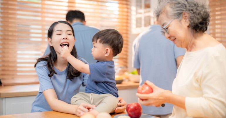 A multi-generational Asian family sits in the kitchen; the grandmother is cutting fruit, and the child feeds it to the mom.