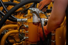 A close-up view shows a person wearing yellow safety gloves working on a yellow hydraulic pump with black hoses.