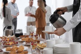 A close-up of a waitress pouring a hot drink during coffee break. The table is full of pastries and mixed fruit.