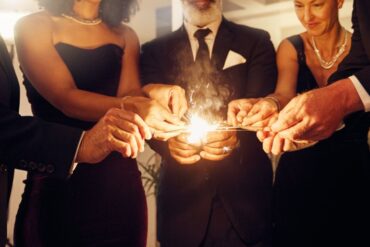 A group of people in black tie attire reaching into the middle of the group and touching lit party sparklers together for fun.