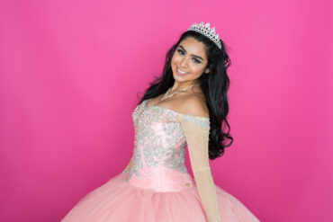 A young woman with dark hair smiles, wearing a sparkly pink off-the-shoulder ball gown and a tiara, against a pink backdrop.