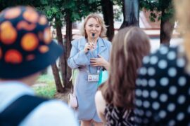 A female tour guide in a blue coat uses a small, handheld microphone to speak to a group of people outdoors.