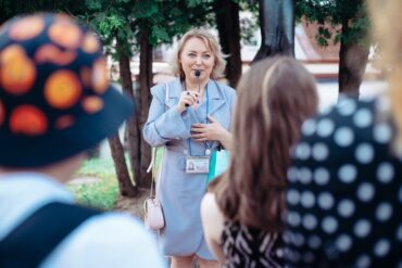 A female tour guide in a blue coat uses a small, handheld microphone to speak to a group of people outdoors.