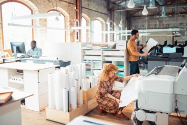 Three people stand in an open concept business office. A man and a woman stand at a printer, and a man sits at a desk.