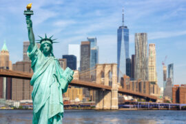 The Statue of Liberty stands tall under the sunlight. Behind the statue is the Manhattan skyline and Brooklyn Bridge.