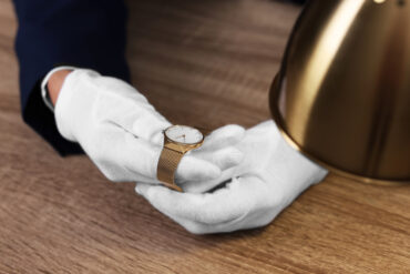 Close-up of two hands wearing white gloves, holding a gold watch under a bright light on top of a wooden surface.