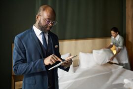 A man in a business suit is using a tablet in a luxury hotel while a young housekeeper is making the bed in the back.