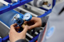 A close-up view shows a person hooking up a gas cylinder to a brass and blue calibration machine.