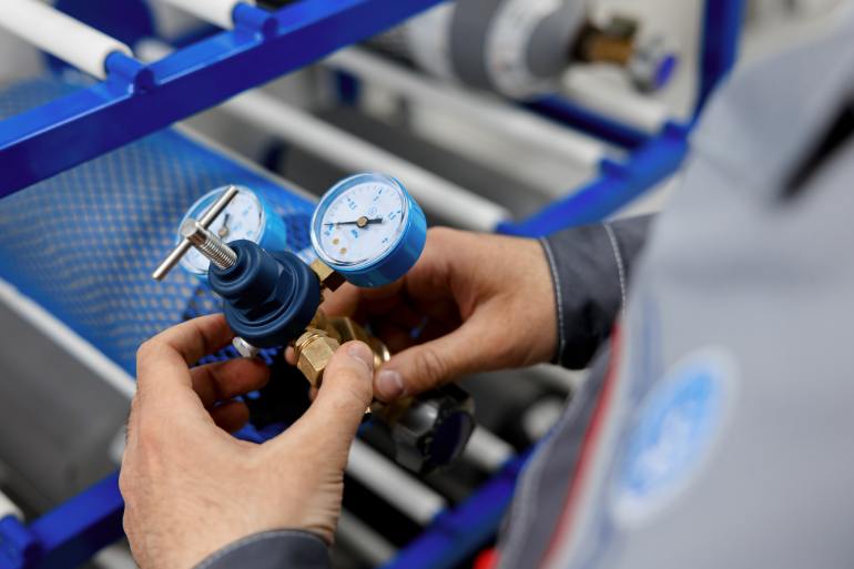 A close-up view shows a person hooking up a gas cylinder to a brass and blue calibration machine.