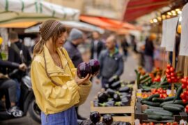 A woman in a yellow jacket and beige head bandana selects an eggplant at a vibrant street market in another country.
