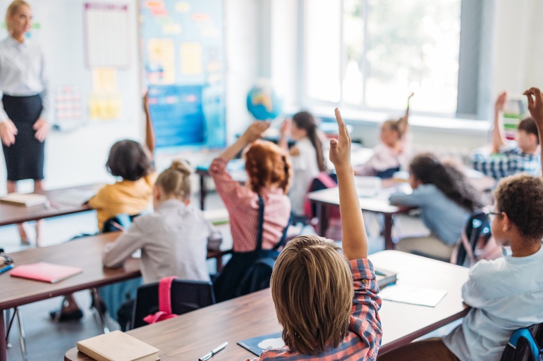 A classroom full of students sitting at their desks. Most students are raising their hands while some are not.