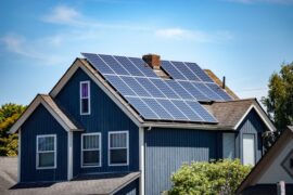 Large blue two-story home with a large angled roof covered in multiple rows of solar panels with a sunny sky.