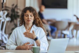 A woman takes a moment to practice mindful breathing while she sits at a table. Her laptop and coffee mug sit on the table.