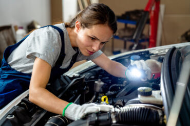 A woman works on a car's engine while holding a small light so she can see. She wears blue overalls.