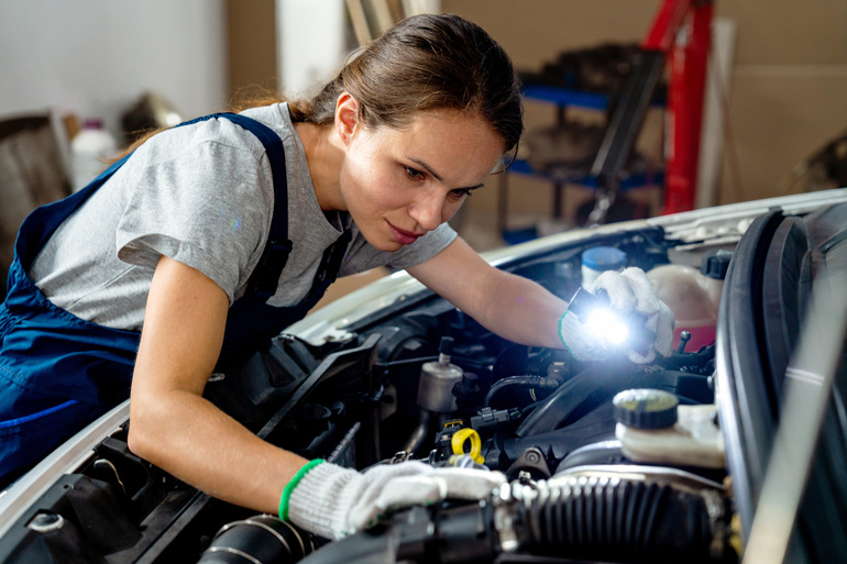 A woman works on a car's engine while holding a small light so she can see. She wears blue overalls.