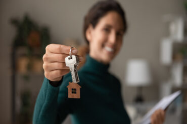 A close-up view shows a woman holding out a set of silver keys with a wooden house cutout hanging on a keychain.