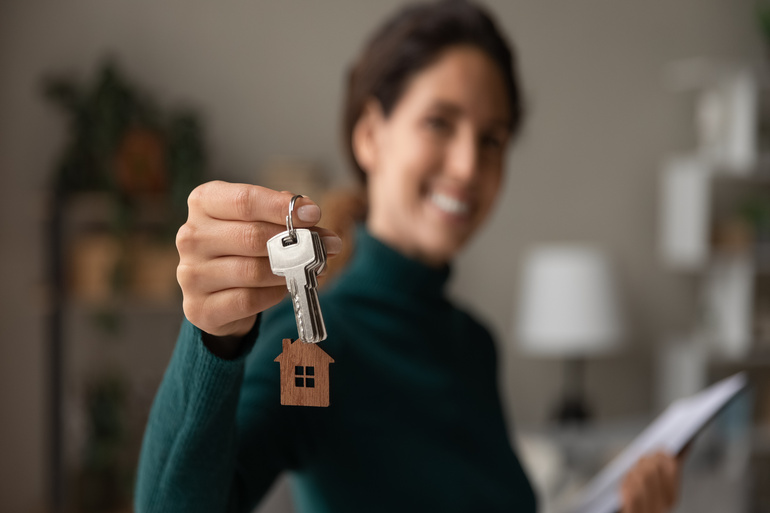 A close-up view shows a woman holding out a set of silver keys with a wooden house cutout hanging on a keychain.