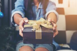 A close-up view shows a woman wearing a denim shirt and jeans holding out a black gift box that's wrapped in a gold ribbon.