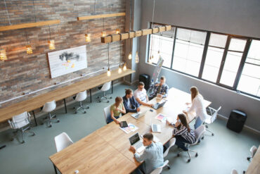 An aerial view of a group of people working at a long wooden conference table in a spacious room with natural lighting.
