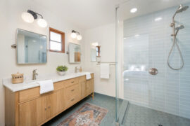 A modern bathroom features a wooden vanity and a walk-in shower with light-blue tile. Two square mirrors hang above the vanity.