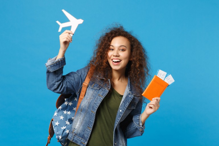 Young woman student in denim clothes, backpack, holding two plane tickets, a plane toy, and an orange passport.