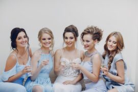 A smiling bride sits alongside her four bridesmaids, in light blue dresses, as they hold glasses of champagne.