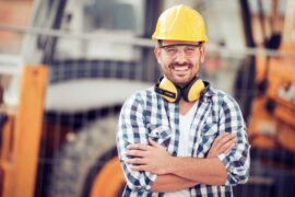 A man wearing a blue plaid shirt, a yellow hard hat and protective glasses smiles with his arms crossed.