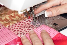 A close-up of an individual using a needle and thread to sew red and pink patterned fabric on a sewing machine.