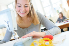 A close-up of a young woman wearing a grey sweater using a sewing machine on a desk, inside a room with more people.