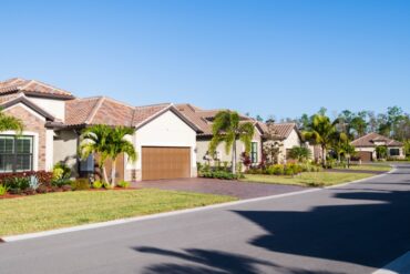 A residential road contains several new houses with brick driveways and brown roofs, palm trees, and grassy lawns.