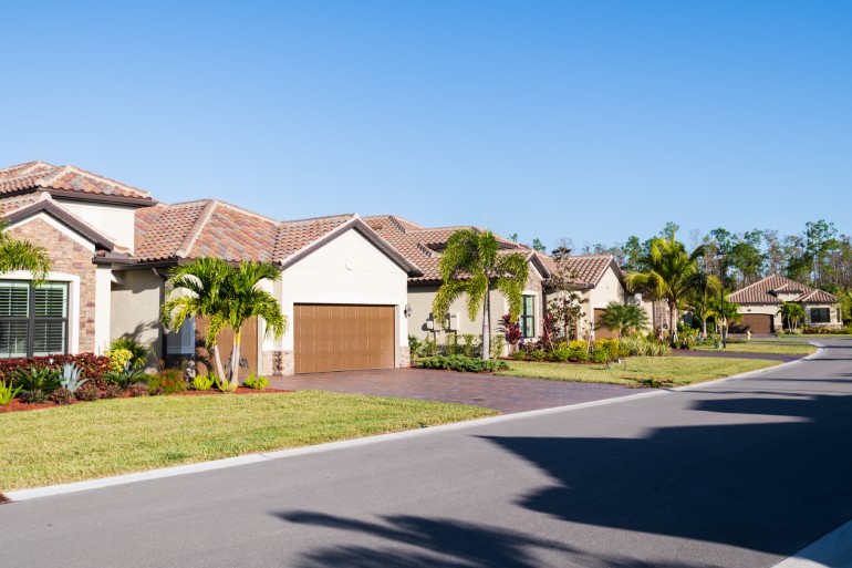 A residential road contains several new houses with brick driveways and brown roofs, palm trees, and grassy lawns.