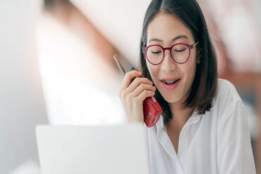 A young woman in red glasses and a white button-down shirt uses a red two-way radio while sitting in front of her laptop.