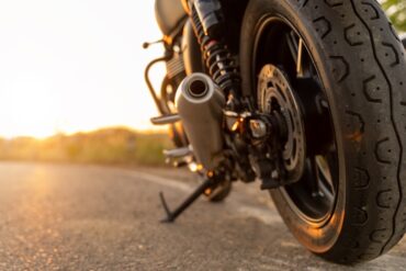 A close-up of a motorcycle's back tire sitting on the road. The kickstand is out and the sun is setting in the back.