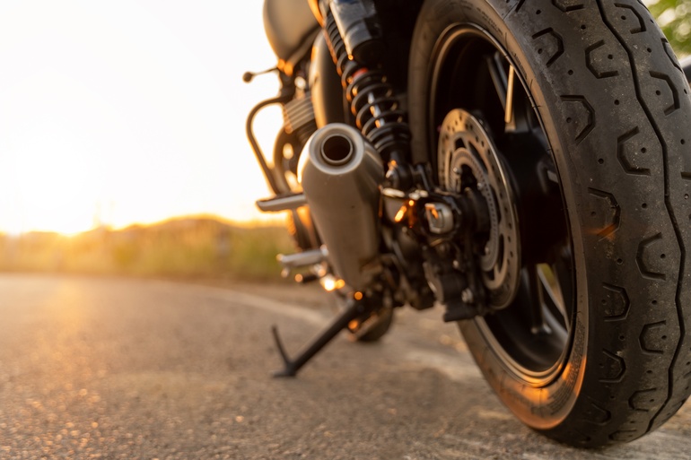 A close-up of a motorcycle's back tire sitting on the road. The kickstand is out and the sun is setting in the back.