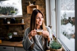 A brunette woman stands next to her kitchen window holding a cup of freshly brewed coffee. She wears a gray bathrobe.