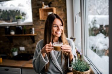 A brunette woman stands next to her kitchen window holding a cup of freshly brewed coffee. She wears a gray bathrobe.