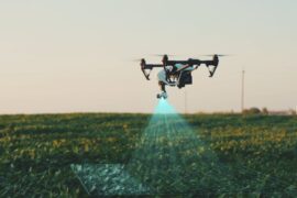 A white agricultural drone flies over the farm fields, scanning and analyzing crops, under a clear sky.